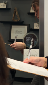 Savoye Salon Spa in Green Bay, WI - A person holds a pen and clipboard near a mirror, reflected while writing—perhaps preparing for their first haircut appointment in Green Bay. A circular sign nearby reads, “ASK FOR AN ABUNDANT TODAY.” The background shows framed documents on shelves, suggesting the careful attention to detail you might expect during a haircut consultation in Green Bay.