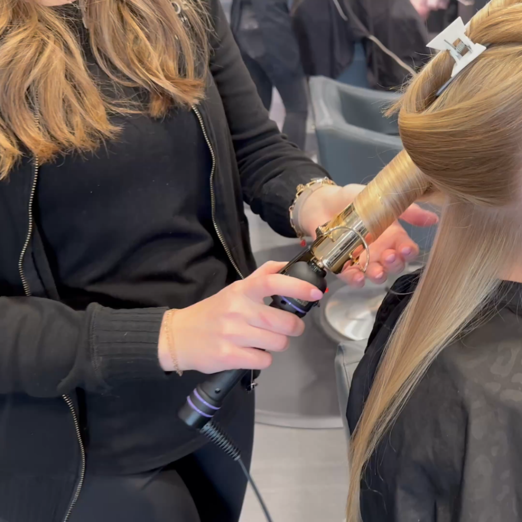 Savoye Salon Spa in Green Bay, WI - A hairstylist uses a curling iron to curl a section of long blonde hair in a salon during a first haircut appointment Green Bay.