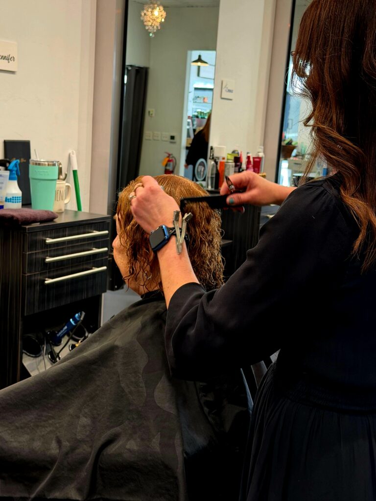 Savoye Salon Spa in Green Bay, WI - A hairstylist combs and trims the wet hair of a seated client in a salon, with various hair products visible on the counter—perfect for those looking to refresh hair between washes or enhance their morning hair routine.